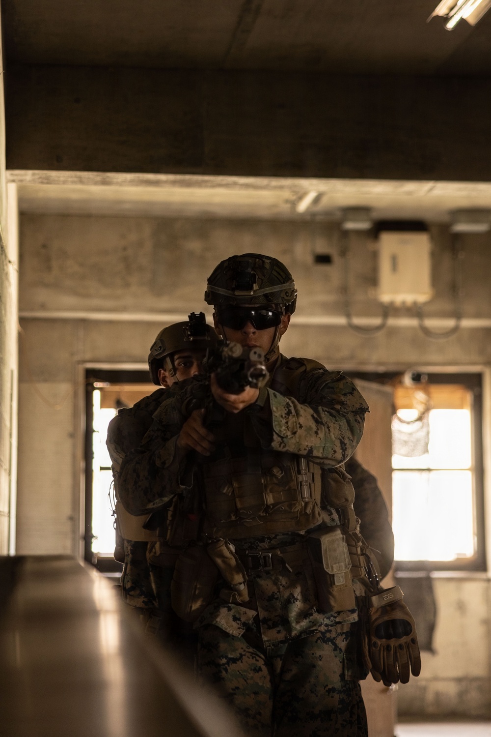 Two military personnel in full combat gear are conducting what appears to be a Military Operations in Urban Terrain (MOUT) training exercise inside a dilapidated concrete building. The lead soldier is aiming a rifle directly toward the camera while the second soldier follows closely behind in a stacked formation. The digital camouflage pattern and equipment are consistent with United States Marine Corps (USMC) MARPAT uniforms.