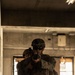 Two military personnel in full combat gear are conducting what appears to be a Military Operations in Urban Terrain (MOUT) training exercise inside a dilapidated concrete building. The lead soldier is aiming a rifle directly toward the camera while the second soldier follows closely behind in a stacked formation. The digital camouflage pattern and equipment are consistent with United States Marine Corps (USMC) MARPAT uniforms.