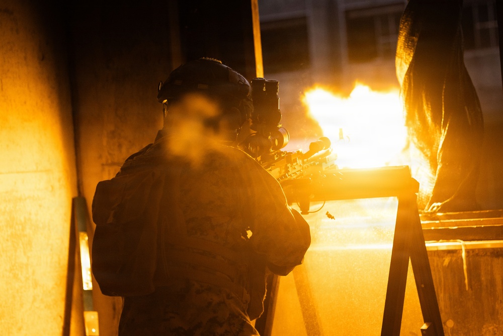 A military or tactical operator is photographed from behind in low-light conditions, aiming a rifle with an optic sight. A large muzzle flash or explosion of flame is visible directly in front of the weapon, illuminating the scene in intense orange light. The operator is wearing a tactical helmet with mounted equipment and camouflage gear.
