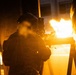 A military or tactical operator is photographed from behind in low-light conditions, aiming a rifle with an optic sight. A large muzzle flash or explosion of flame is visible directly in front of the weapon, illuminating the scene in intense orange light. The operator is wearing a tactical helmet with mounted equipment and camouflage gear.