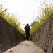 A lone figure wearing dark clothing, a bucket hat, and carrying a backpack walks away from the camera along a narrow concrete pathway flanked by two weathered stone or concrete walls. Lush green vegetation and tall grasses grow above the walls on both sides, framing the scene. The composition uses strong linear perspective, drawing the eye toward the silhouetted subject against a bright, overcast sky.