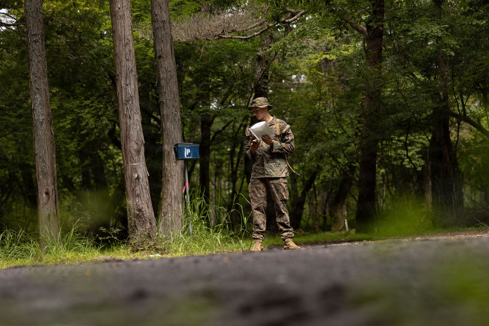 A military service member in camouflage uniform and boonie hat stands on a path in a wooded area, reviewing documents or papers. The setting appears to be a training area or field exercise location, indicated by a blue marker post visible near the trees. The image is taken from a low angle with a shallow depth of field.