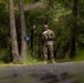 A military service member in camouflage uniform and boonie hat stands on a path in a wooded area, reviewing documents or papers. The setting appears to be a training area or field exercise location, indicated by a blue marker post visible near the trees. The image is taken from a low angle with a shallow depth of field.