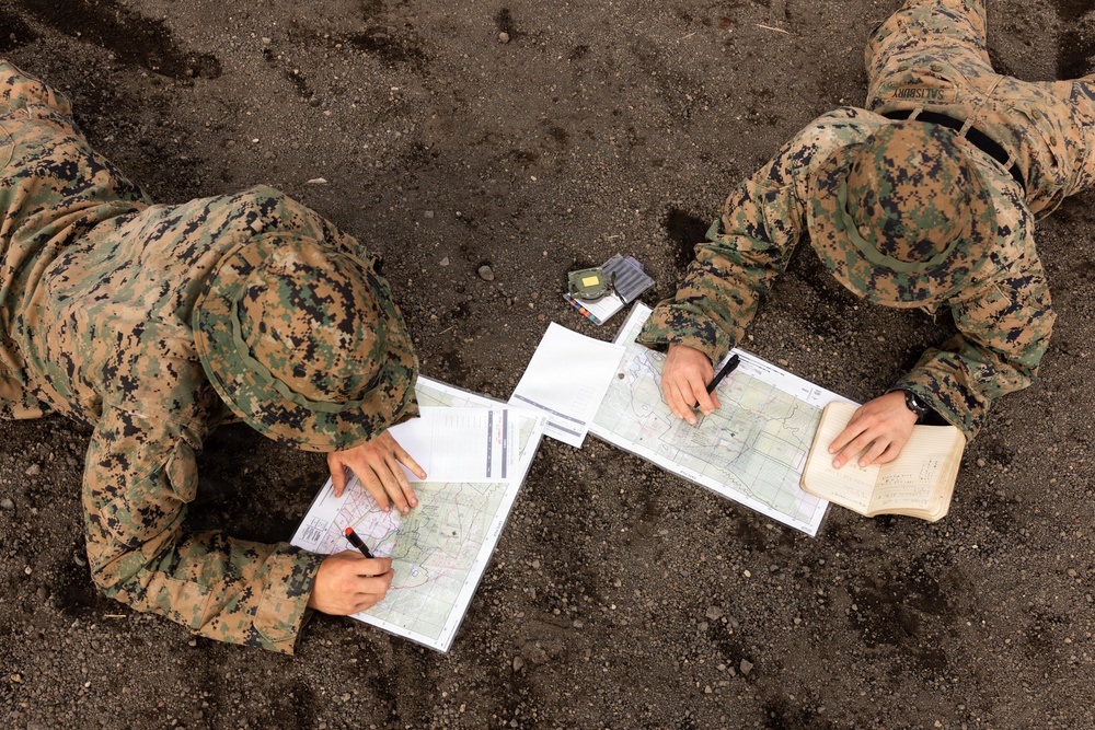 Two military personnel in MARPAT digital camouflage uniforms are lying prone on bare ground, conducting map reading and land navigation exercises. They are working with topographic maps, writing instruments, and what appears to be a compass or small device placed between them. The name tape 'SALISBURY' is visible on the right individual's uniform.