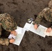 Two military personnel in MARPAT digital camouflage uniforms are lying prone on bare ground, conducting map reading and land navigation exercises. They are working with topographic maps, writing instruments, and what appears to be a compass or small device placed between them. The name tape 'SALISBURY' is visible on the right individual's uniform.