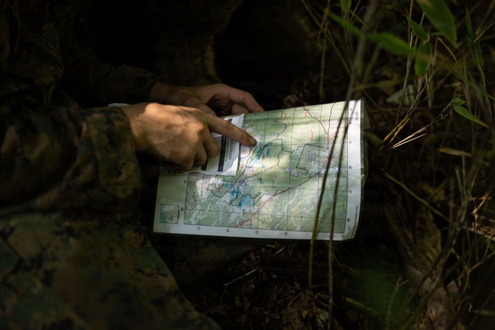 A person wearing military camouflage (MARPAT pattern consistent with U.S. Marines) is examining a topographic map in a dense jungle or forest environment. The individual is pointing at a specific location on the map, suggesting navigation or route planning. The setting appears to be a field training or operational environment with low ambient light.