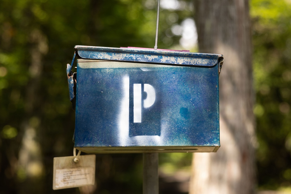 The image shows a weathered blue metal box, likely a repurposed ammunition can or utility container, suspended by a wire cable. A white stenciled letter 'P' is painted on its front face, and a small paper tag hangs from the bottom. The box appears to be used as a makeshift mailbox or information holder, mounted near a wooden post in an outdoor, tree-lined setting.