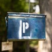 The image shows a weathered blue metal box, likely a repurposed ammunition can or utility container, suspended by a wire cable. A white stenciled letter 'P' is painted on its front face, and a small paper tag hangs from the bottom. The box appears to be used as a makeshift mailbox or information holder, mounted near a wooden post in an outdoor, tree-lined setting.