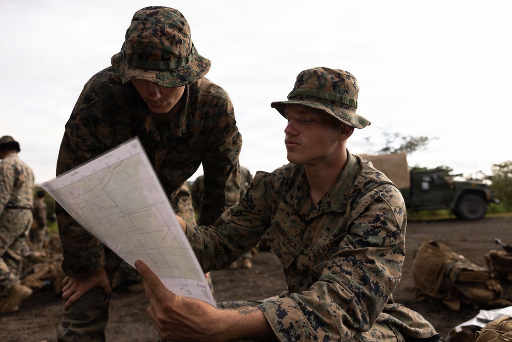 Two U.S. Marines in MARPAT woodland camouflage uniforms and boonie covers are reviewing a topographic map together in a field environment. The foreground Marine's uniform displays 'U.S. MARINES' tape on the chest. Additional Marines and a military vehicle are visible in the background.