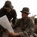 Two U.S. Marines in MARPAT woodland camouflage uniforms and boonie covers are reviewing a topographic map together in a field environment. The foreground Marine's uniform displays 'U.S. MARINES' tape on the chest. Additional Marines and a military vehicle are visible in the background.