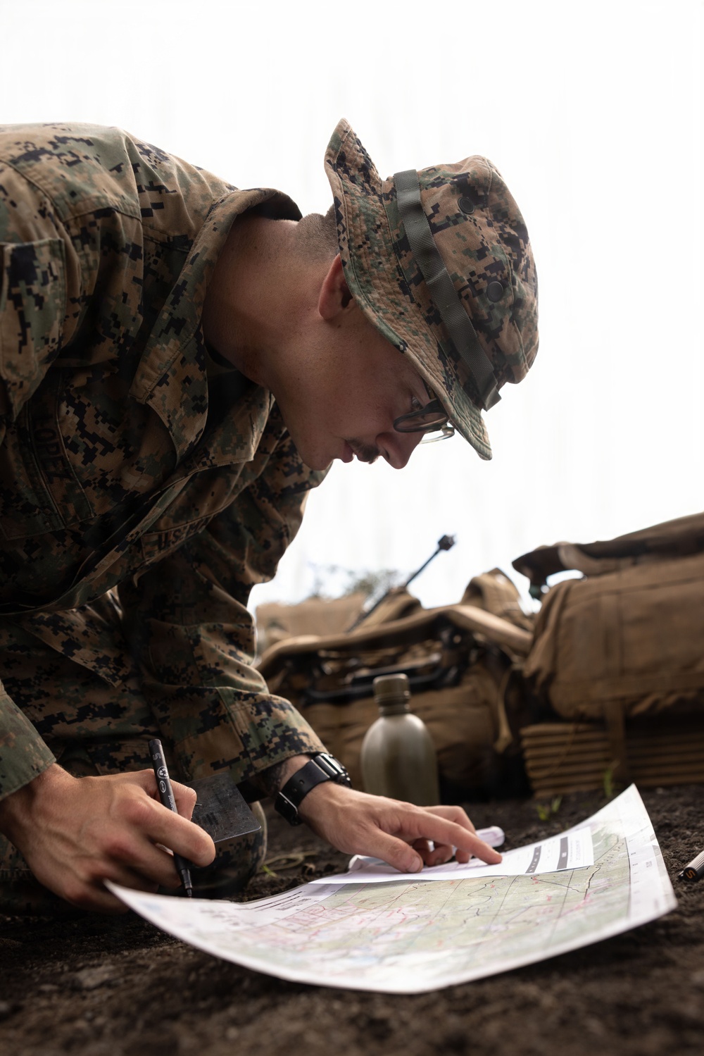 A U.S. Marine in MARPAT digital camouflage uniform and boonie cover is leaning over a military topographic map, marking or annotating it with a pen. The setting appears to be a field environment with military gear visible in the background. The image captures a map-reading or mission-planning activity in an operational or training context.