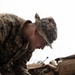 A U.S. Marine in MARPAT digital camouflage uniform and boonie cover is leaning over a military topographic map, marking or annotating it with a pen. The setting appears to be a field environment with military gear visible in the background. The image captures a map-reading or mission-planning activity in an operational or training context.