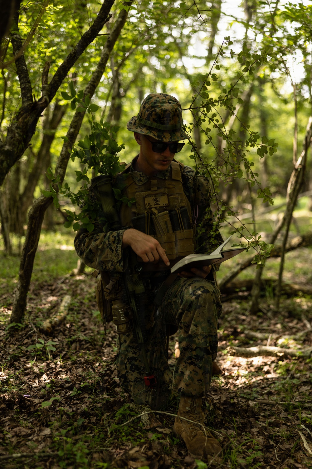 A military service member in full camouflage gear stands in a wooded area reviewing what appears to be a map or notebook. The individual is wearing MARPAT (Marine pattern) digital camouflage uniform and tactical vest, suggesting U.S. Marine Corps affiliation. The setting appears to be a training exercise or field operation in a deciduous forest environment.