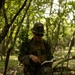 A military service member in full camouflage gear stands in a wooded area reviewing what appears to be a map or notebook. The individual is wearing MARPAT (Marine pattern) digital camouflage uniform and tactical vest, suggesting U.S. Marine Corps affiliation. The setting appears to be a training exercise or field operation in a deciduous forest environment.