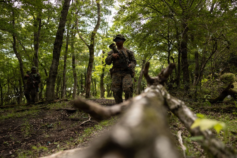 A military patrol moves through a dense, green forest along a dirt trail. The primary subject is a soldier in woodland camouflage uniform and boonie hat, carrying a rifle at the ready. At least one additional soldier is visible in the background to the left, suggesting a small unit patrol formation.