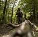 A military patrol moves through a dense, green forest along a dirt trail. The primary subject is a soldier in woodland camouflage uniform and boonie hat, carrying a rifle at the ready. At least one additional soldier is visible in the background to the left, suggesting a small unit patrol formation.