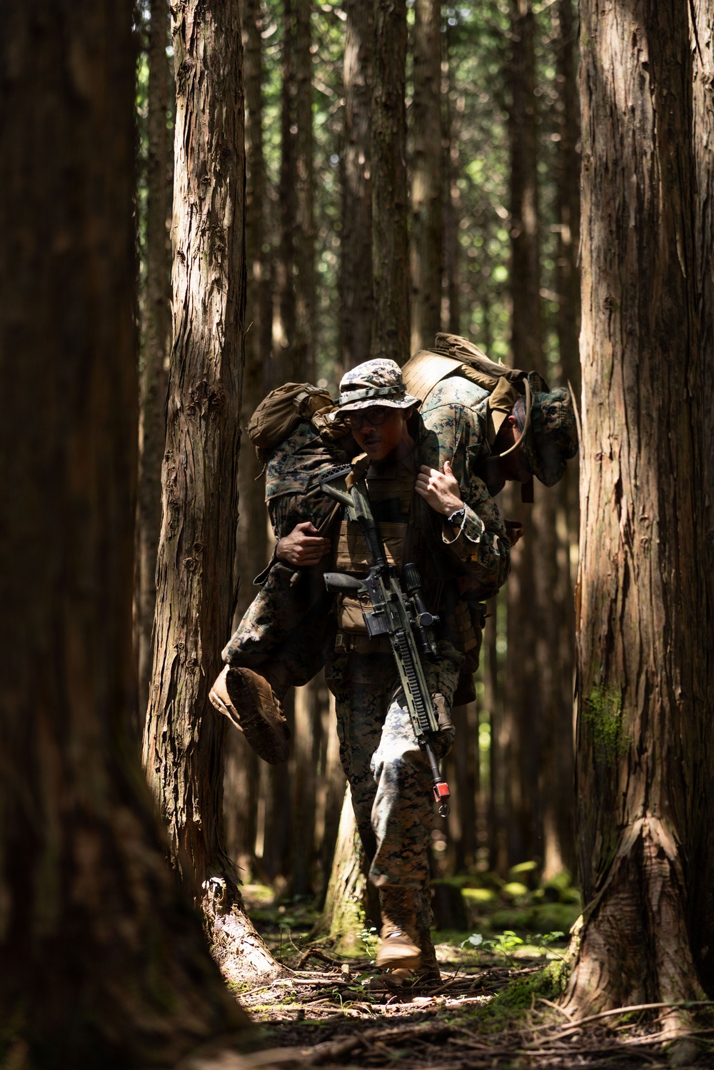 A U.S. Marine in MARPAT woodland camouflage is performing a fireman's carry, transporting an incapacitated fellow Marine through a dense forest. The carrying Marine is armed with an M4-style carbine and wearing a boonie cover, tactical vest, and full combat load. The setting appears to be a training exercise in a tall-canopy forest environment.