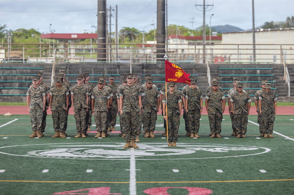 A formation of U.S. Marines in woodland MARPAT camouflage uniforms stands at attention on a green turf football field. A red guidon flag is visible in the center of the formation, bearing USMC insignia and partial unit designation text. The setting appears to be a military base athletic field with stadium bleachers and tropical vegetation visible in the background.