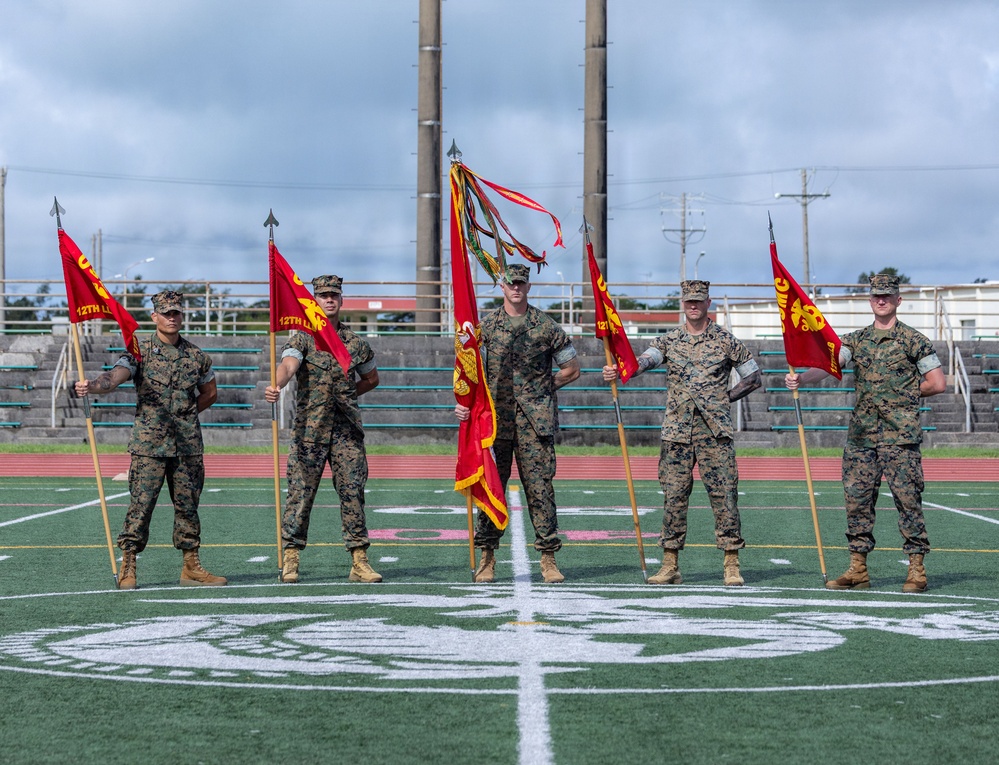 Five U.S. Marines in woodland MARPAT camouflage uniforms stand in formation on a military athletic field, holding unit guidons and a regimental or battalion colors. The flags are red with yellow markings and include text referencing '12th Marines,' suggesting this is a formal military ceremony such as a change of command or activation event. The setting appears to be a military installation in a tropical or subtropical environment, likely in the Pacific region.