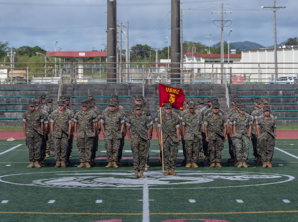 A formation of U.S. Marines stands at attention on an athletic field, with a guidon bearer holding a red USMC pennant flag at the center front. The Marines are wearing woodland MARPAT camouflage uniforms and tan combat boots, suggesting a formal military ceremony or muster. The setting appears to be a military base athletic facility with bleachers and utility infrastructure visible in the background.