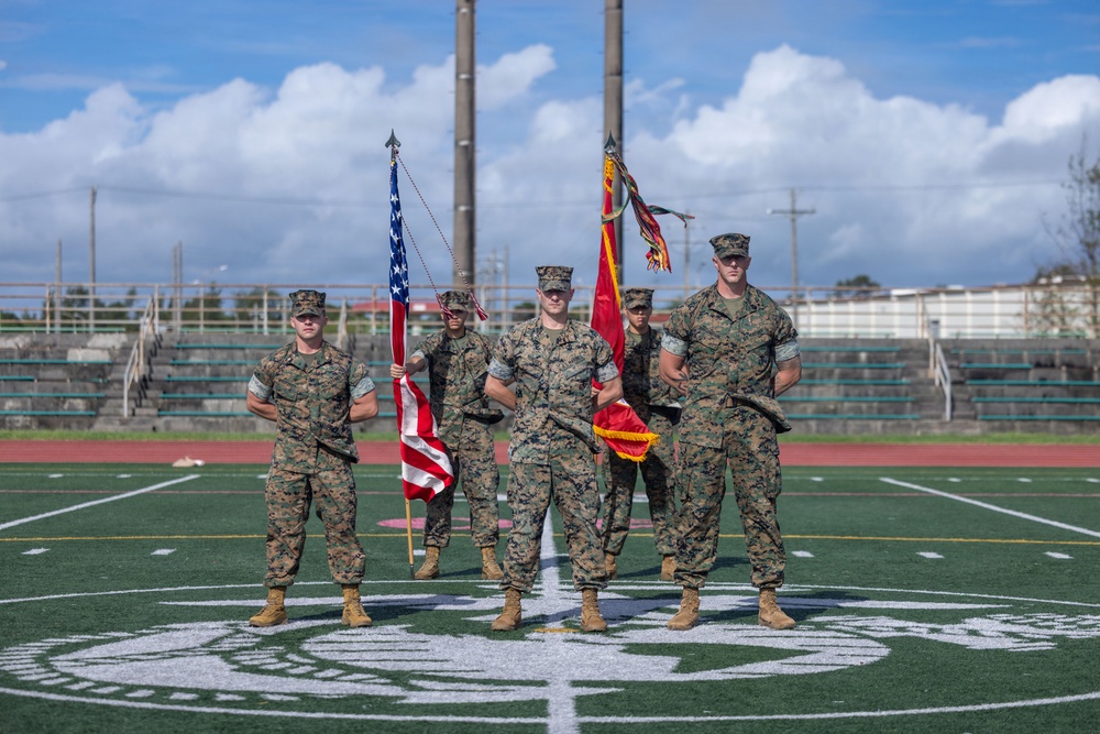 Five U.S. Marine Corps personnel in MARPAT camouflage uniforms stand in formation on a football field, holding the American flag and what appears to be a unit guidon or colors. The setting appears to be a formal military ceremony, likely a change of command or similar official event. The field displays a Marine Corps emblem painted on the turf, and stadium bleachers are visible in the background.