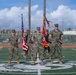 Five U.S. Marine Corps personnel in MARPAT camouflage uniforms stand in formation on a football field, holding the American flag and what appears to be a unit guidon or colors. The setting appears to be a formal military ceremony, likely a change of command or similar official event. The field displays a Marine Corps emblem painted on the turf, and stadium bleachers are visible in the background.