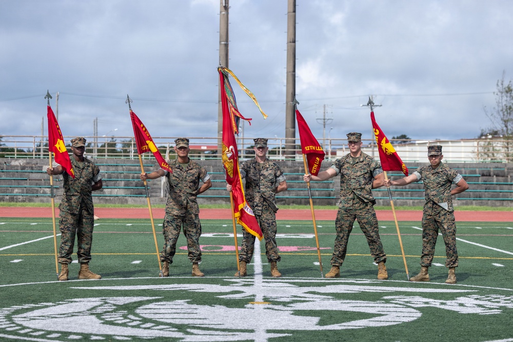 Six U.S. Marines in woodland MARPAT camouflage uniforms stand in formation on a football field holding red guidons and unit colors. The flags visible include what appear to be 12th Marines regiment colors and subordinate unit guidons. The setting appears to be a military ceremony on an athletic field, likely at a base in the Pacific region based on the surroundings.