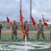 Six U.S. Marines in woodland MARPAT camouflage uniforms stand in formation on a football field holding red guidons and unit colors. The flags visible include what appear to be 12th Marines regiment colors and subordinate unit guidons. The setting appears to be a military ceremony on an athletic field, likely at a base in the Pacific region based on the surroundings.