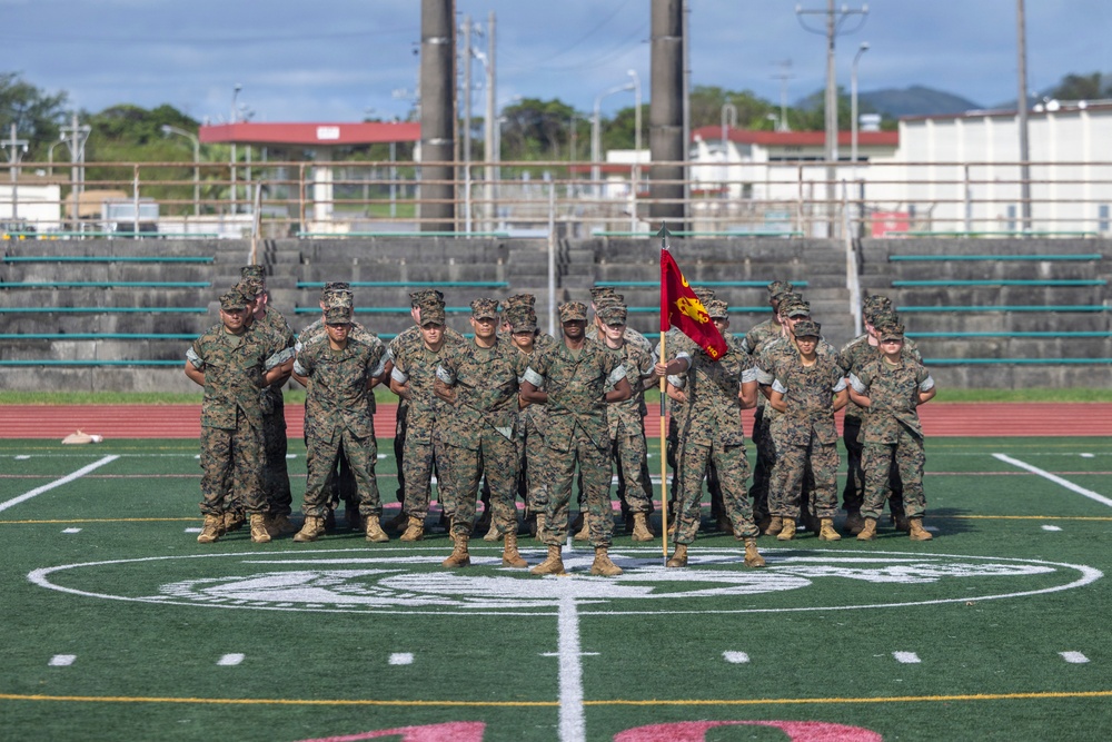 A group of U.S. Marines in MARPAT camouflage uniforms stand in formation on a green turf football field, with one Marine holding a red unit guidon flag. The field displays a Marine Corps emblem at center field, suggesting this is a military base athletic facility. The setting appears to be a formal unit gathering or physical training formation event.