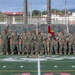 A group of U.S. Marines in MARPAT camouflage uniforms stand in formation on a green turf football field, with one Marine holding a red unit guidon flag. The field displays a Marine Corps emblem at center field, suggesting this is a military base athletic facility. The setting appears to be a formal unit gathering or physical training formation event.