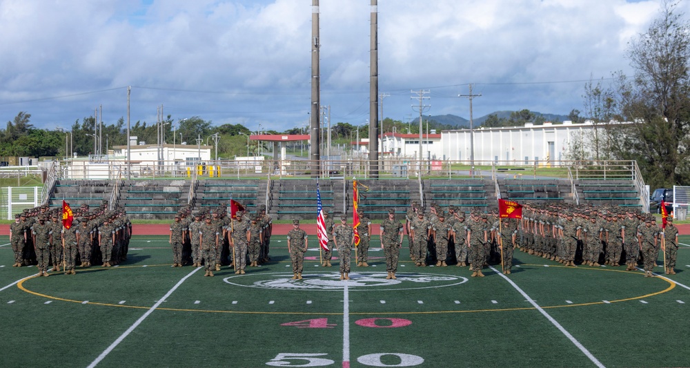 A large formation of U.S. Marines in woodland MARPAT camouflage uniforms is assembled on a football field at the 50-yard line, likely during a formal ceremony or parade. Multiple unit guidons and the American flag are prominently displayed at the center of the formation. The setting appears to be a military base athletic field with bleachers, industrial structures, and tropical vegetation visible in the background.