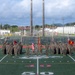 A large formation of U.S. Marines in woodland MARPAT camouflage uniforms is assembled on a football field at the 50-yard line, likely during a formal ceremony or parade. Multiple unit guidons and the American flag are prominently displayed at the center of the formation. The setting appears to be a military base athletic field with bleachers, industrial structures, and tropical vegetation visible in the background.