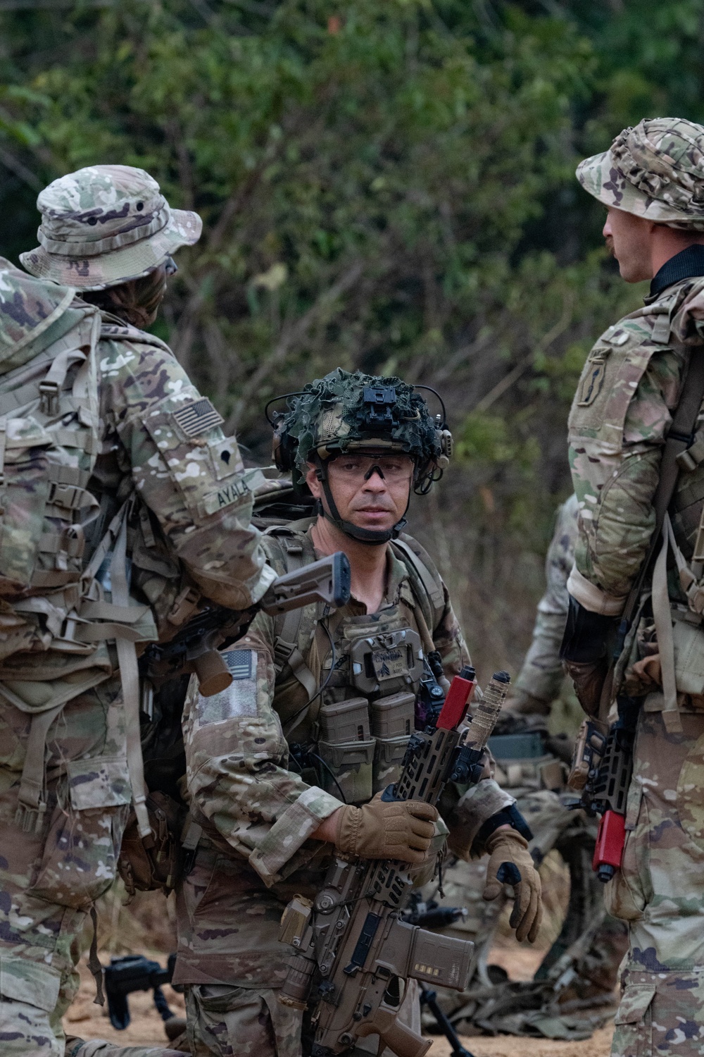 Three military personnel in MultiCam/OCP camouflage uniforms are gathered in what appears to be a field training environment with wooded terrain in the background. The central figure is wearing a tactical helmet with night vision mount and communications headset, holding an assault rifle with a red training/blank-firing adapter. The soldiers appear to be conducting a briefing or coordination during a military exercise.
