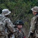 Three military personnel in MultiCam/OCP camouflage uniforms are gathered in what appears to be a field training environment with wooded terrain in the background. The central figure is wearing a tactical helmet with night vision mount and communications headset, holding an assault rifle with a red training/blank-firing adapter. The soldiers appear to be conducting a briefing or coordination during a military exercise.
