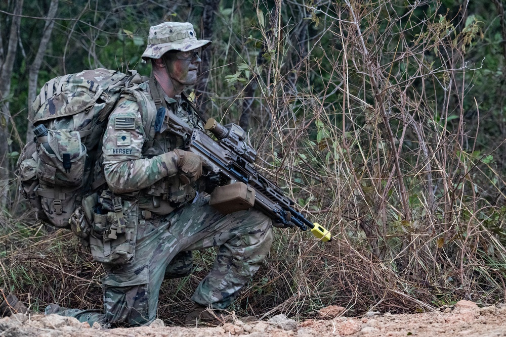 A U.S. Army soldier identified by name tape as 'HERSEY' is in a kneeling tactical position in a wooded outdoor environment. The soldier is wearing full MultiCam (OCP) camouflage uniform with face paint applied, carrying a large rucksack, and holding what appears to be an M249 SAW or similar light machine gun with a yellow training/safety device attached to the muzzle. The yellow muzzle device and MILES-type gear suggest this is a field training exercise rather than a combat deployment.