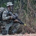 A U.S. Army soldier identified by name tape as 'HERSEY' is in a kneeling tactical position in a wooded outdoor environment. The soldier is wearing full MultiCam (OCP) camouflage uniform with face paint applied, carrying a large rucksack, and holding what appears to be an M249 SAW or similar light machine gun with a yellow training/safety device attached to the muzzle. The yellow muzzle device and MILES-type gear suggest this is a field training exercise rather than a combat deployment.