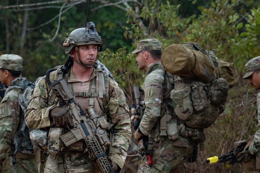 A group of military personnel in camouflage uniforms are gathered in a wooded outdoor environment, appearing to be conducting a field training exercise or tactical operation. The foreground soldier is heavily equipped with full combat gear including a tactical helmet with night vision mount, body armor, tactical vest, and an assault rifle. Multiple soldiers are visible in the background, some carrying large rucksacks.
