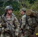A group of military personnel in camouflage uniforms are gathered in a wooded outdoor environment, appearing to be conducting a field training exercise or tactical operation. The foreground soldier is heavily equipped with full combat gear including a tactical helmet with night vision mount, body armor, tactical vest, and an assault rifle. Multiple soldiers are visible in the background, some carrying large rucksacks.
