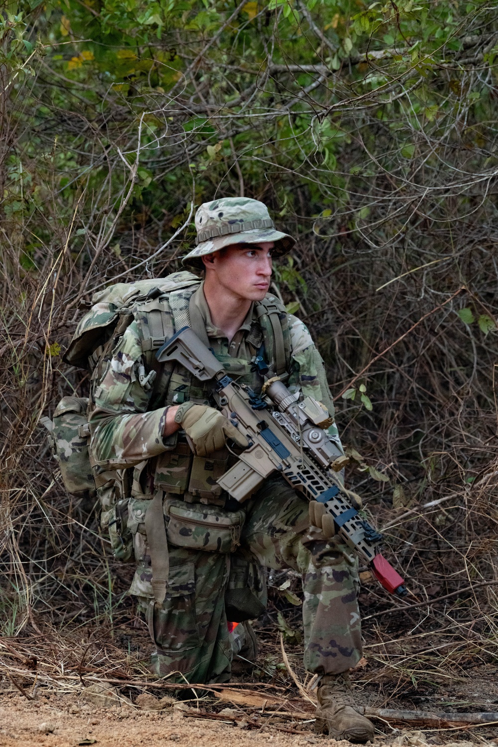 A young male soldier in full combat gear is positioned in a wooded/brushy environment, appearing to be on patrol or a field training exercise. He is equipped with multicam camouflage uniform, a boonie hat, a large rucksack, and is carrying what appears to be an M4/AR-platform rifle with optics and accessories. A red training/safety flag is visible on the weapon, suggesting this may be a training exercise rather than a live combat situation.