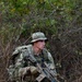 A young male soldier in full combat gear is positioned in a wooded/brushy environment, appearing to be on patrol or a field training exercise. He is equipped with multicam camouflage uniform, a boonie hat, a large rucksack, and is carrying what appears to be an M4/AR-platform rifle with optics and accessories. A red training/safety flag is visible on the weapon, suggesting this may be a training exercise rather than a live combat situation.