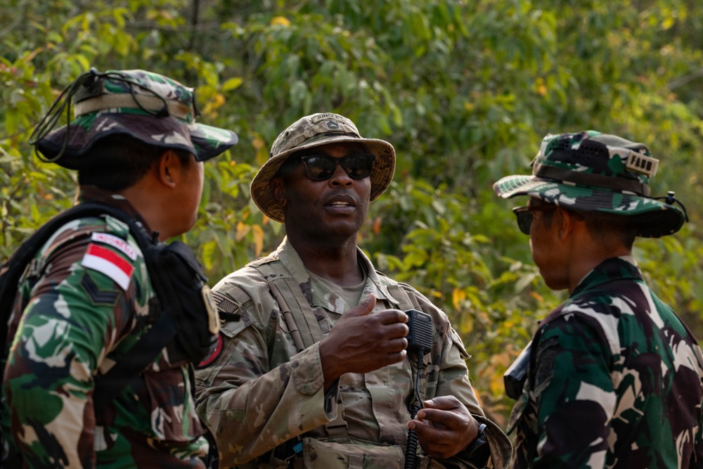 Three military personnel are engaged in a discussion in an outdoor, wooded environment. The central figure wears US Army multicam uniform and sunglasses, while the two flanking individuals wear Indonesian Army (TNI-AD) camouflage uniforms with distinctive Indonesian flag patches. This appears to be a joint military exercise or coordination meeting between US and Indonesian armed forces.