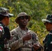 Three military personnel are engaged in a discussion in an outdoor, wooded environment. The central figure wears US Army multicam uniform and sunglasses, while the two flanking individuals wear Indonesian Army (TNI-AD) camouflage uniforms with distinctive Indonesian flag patches. This appears to be a joint military exercise or coordination meeting between US and Indonesian armed forces.