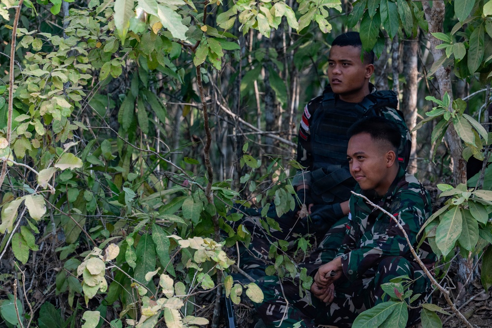 Two young men are positioned in a dense forest or jungle environment. The foreground individual wears military camouflage uniform, while the person behind wears a black tactical vest over a dark shirt. Both appear alert and are observing something off-camera.