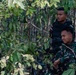 Two young men are positioned in a dense forest or jungle environment. The foreground individual wears military camouflage uniform, while the person behind wears a black tactical vest over a dark shirt. Both appear alert and are observing something off-camera.