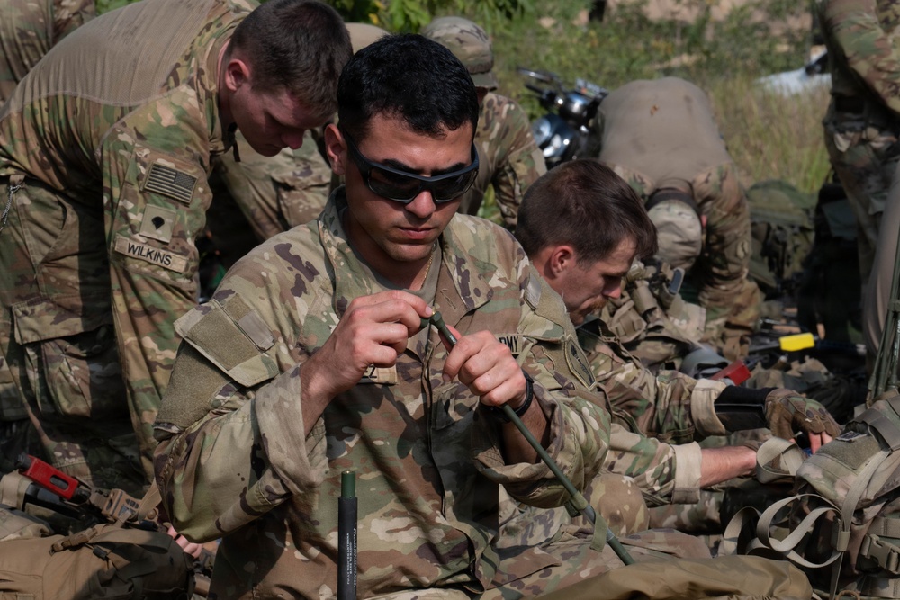 Multiple U.S. Army soldiers in MultiCam/OCP camouflage uniforms are engaged in what appears to be a field training exercise or equipment preparation activity. The central soldier, wearing ballistic eyewear, is focused on handling what appears to be a green cord or detonation cord/demo material. Several soldiers are working simultaneously on similar tasks in an outdoor setting.
