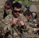 Multiple U.S. Army soldiers in MultiCam/OCP camouflage uniforms are engaged in what appears to be a field training exercise or equipment preparation activity. The central soldier, wearing ballistic eyewear, is focused on handling what appears to be a green cord or detonation cord/demo material. Several soldiers are working simultaneously on similar tasks in an outdoor setting.