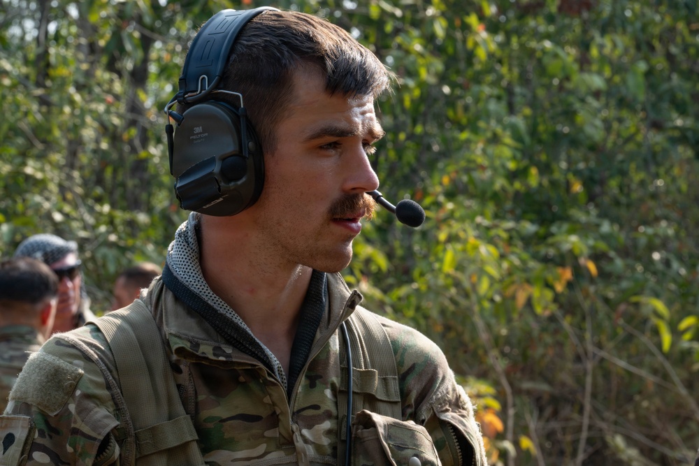 A young male individual wearing military multicam camouflage uniform and a 3M Peltor tactical communications headset with boom microphone is photographed in a outdoor wooded environment. He appears to be actively communicating via the headset while facing to his right. Additional personnel in military attire are partially visible in the background.
