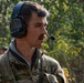 A young male individual wearing military multicam camouflage uniform and a 3M Peltor tactical communications headset with boom microphone is photographed in a outdoor wooded environment. He appears to be actively communicating via the headset while facing to his right. Additional personnel in military attire are partially visible in the background.