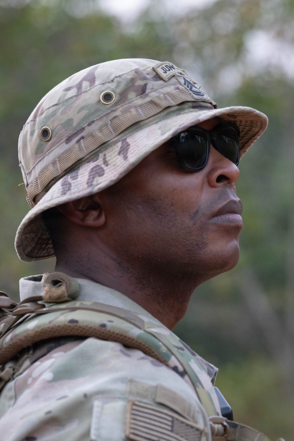 A military service member is photographed in a close-up profile shot, wearing MultiCam/OCP pattern camouflage uniform and boonie hat with a name tape partially visible. The individual is equipped with tactical gear including a backpack/rucksack and is wearing dark sunglasses. The outdoor background shows blurred vegetation, suggesting a field or training environment.