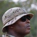 A military service member is photographed in a close-up profile shot, wearing MultiCam/OCP pattern camouflage uniform and boonie hat with a name tape partially visible. The individual is equipped with tactical gear including a backpack/rucksack and is wearing dark sunglasses. The outdoor background shows blurred vegetation, suggesting a field or training environment.
